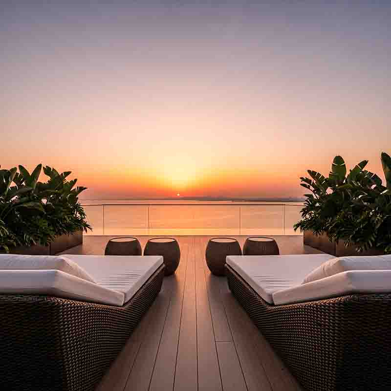 Eine luxuriöse Dachterrasse von Ciel Dubai Marina bei Sonnenuntergang. Die Terrasse verfügt über einen Holzfußboden, zwei dunkle Rattan-Sonnenliegen mit weißen Polstern, flankiert von grünen Pflanzen in Pflanzkübeln, und bietet einen weiten Blick über das Meer von Dubai.
