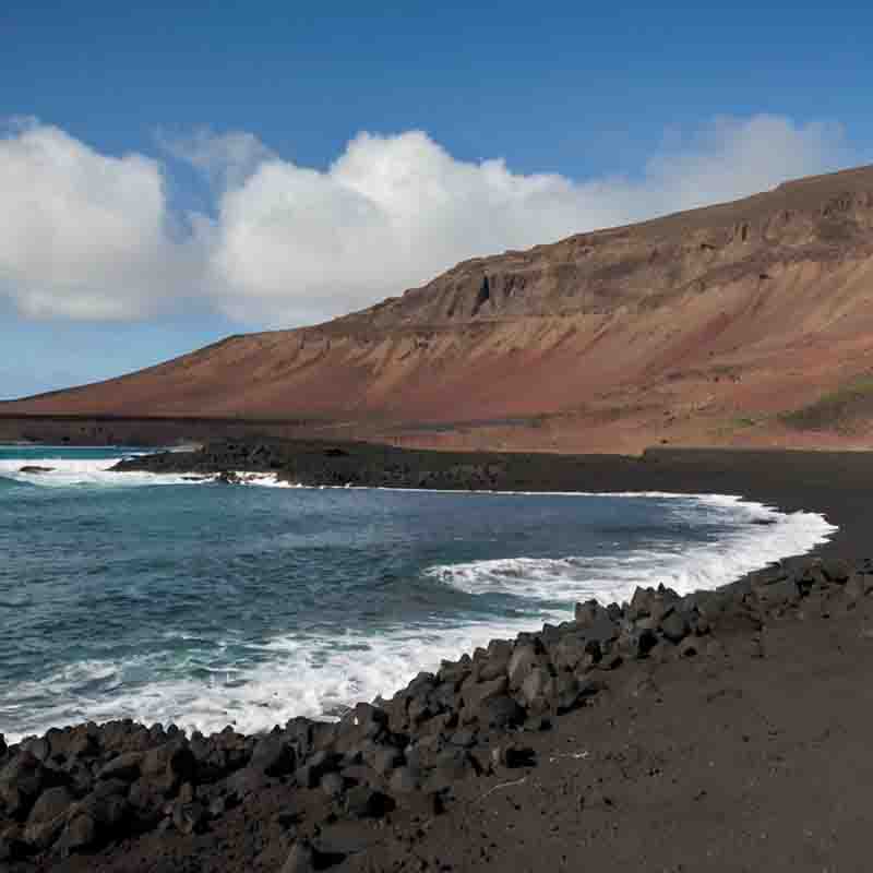 Ein schwarzer Sandstrand auf Lanzarote mit majestätischen Bergen im Hintergrund, der eine beeindruckende natürliche Landschaft zeigt.