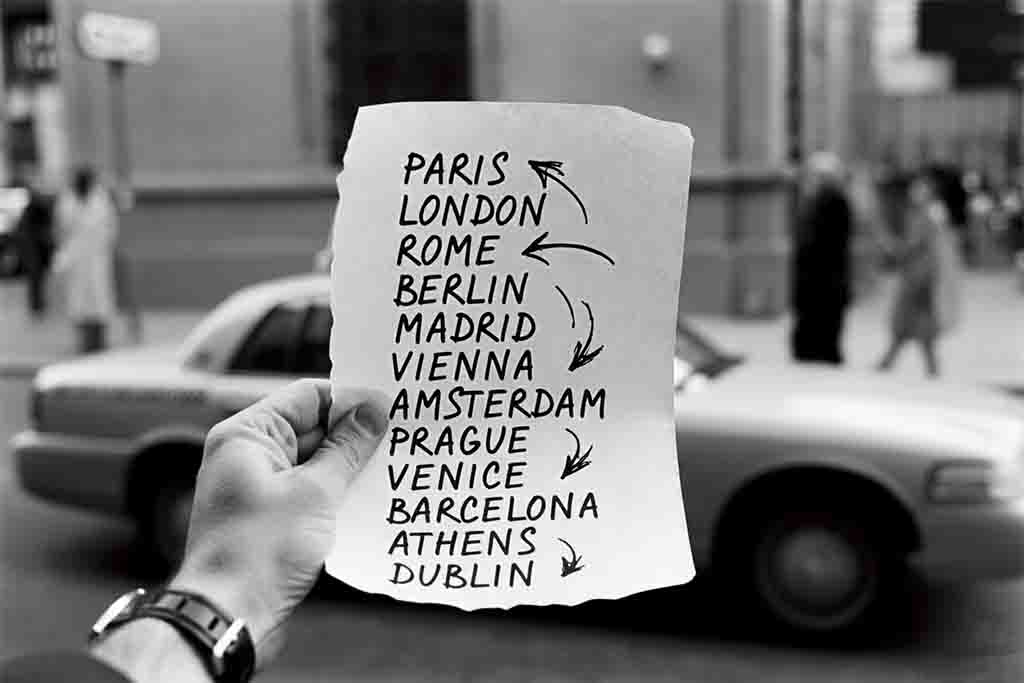 Eine Hand hält einen Zettel mit einer Liste europäischer Städte wie Paris, London und Rom für die Reiseplanung vor einem unscharfen Stadthintergrund mit einem Taxi.