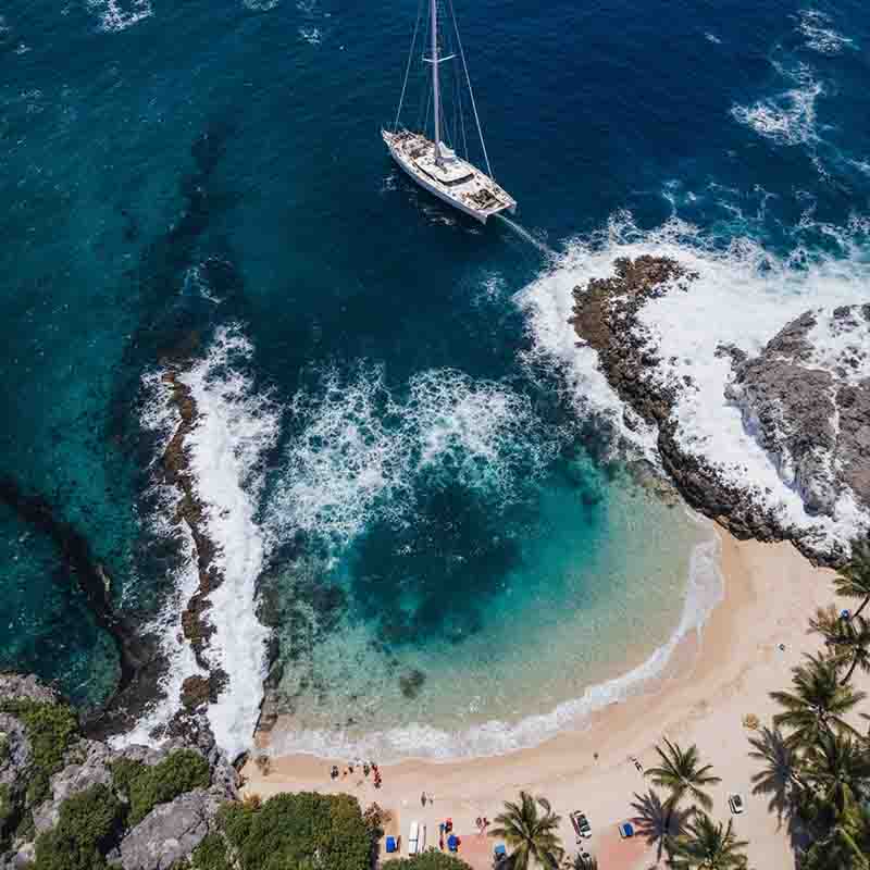 Panoramablick auf einen Segelkatamaran, der in einer kleinen, ruhigen Bucht in Puerto Rico mit türkisfarbenem Wasser, weißem Sandstrand und grünen Palmen vor einer felsigen Küste ankert.